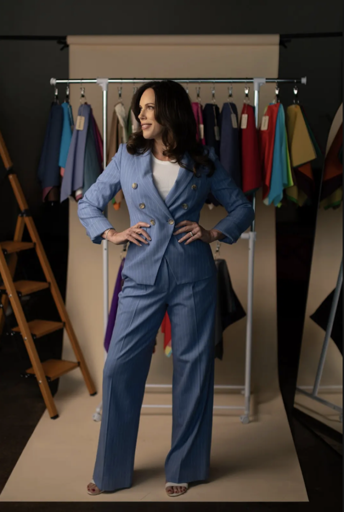 Woman in a blue suit stands confidently in front of a clothing rack with colorful garments, next to a wooden ladder, in a studio setting—an ideal scene for professional headshots Dallas clients would expect from an expert Dallas photographer.