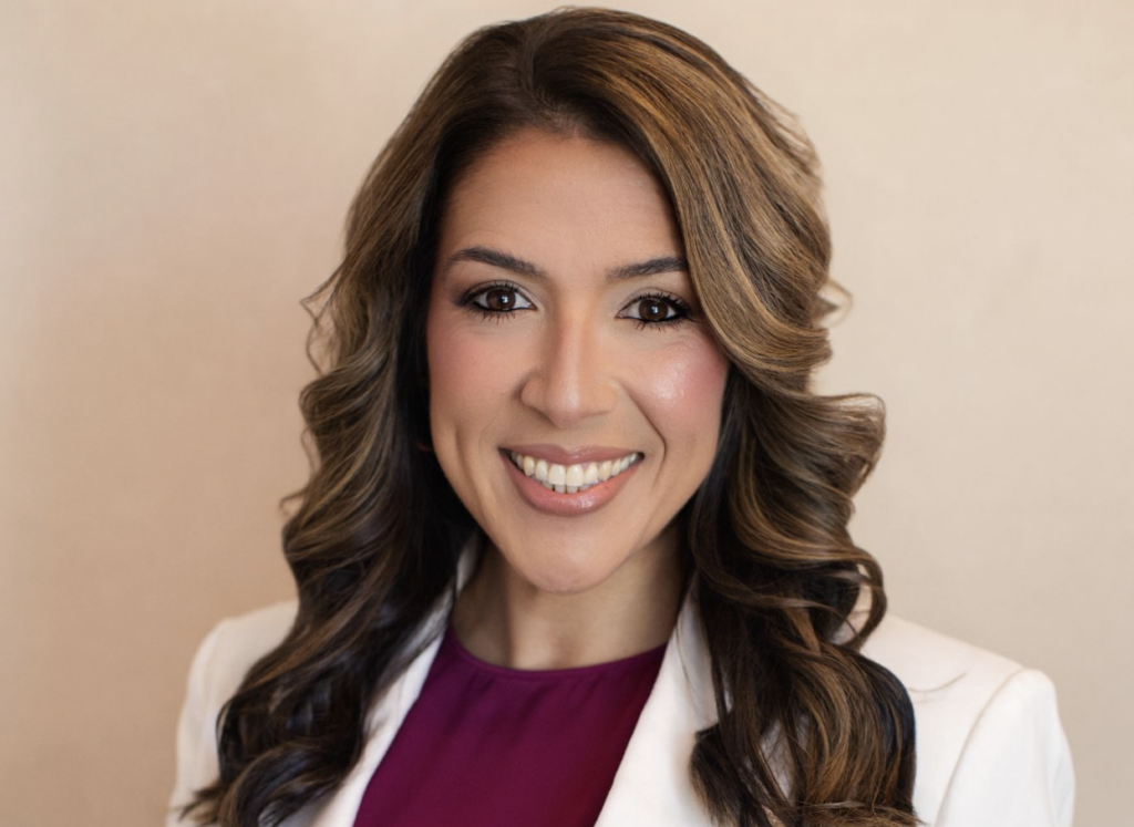 Smiling professional woman with long wavy brown hair wearing a white blazer and burgundy top