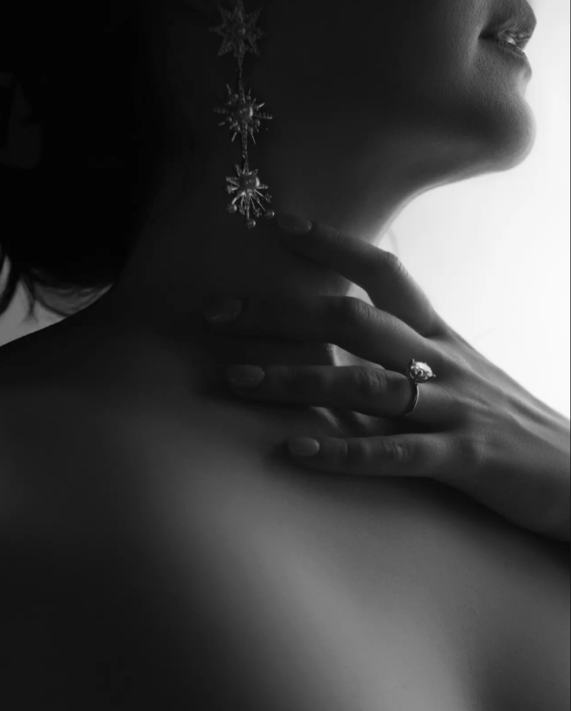 A woman’s hand with a ring rests on her collarbone. She wears long, star-shaped earrings. The image is in black and white with soft lighting, perfectly capturing the intimate allure of Boudoir Sessions.