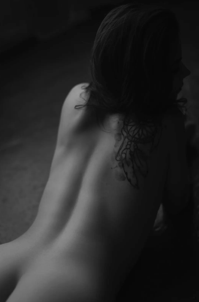 A person lies on their stomach, photographed from behind in black and white, showcasing back and shoulder tattoos during a boudoir photography session.