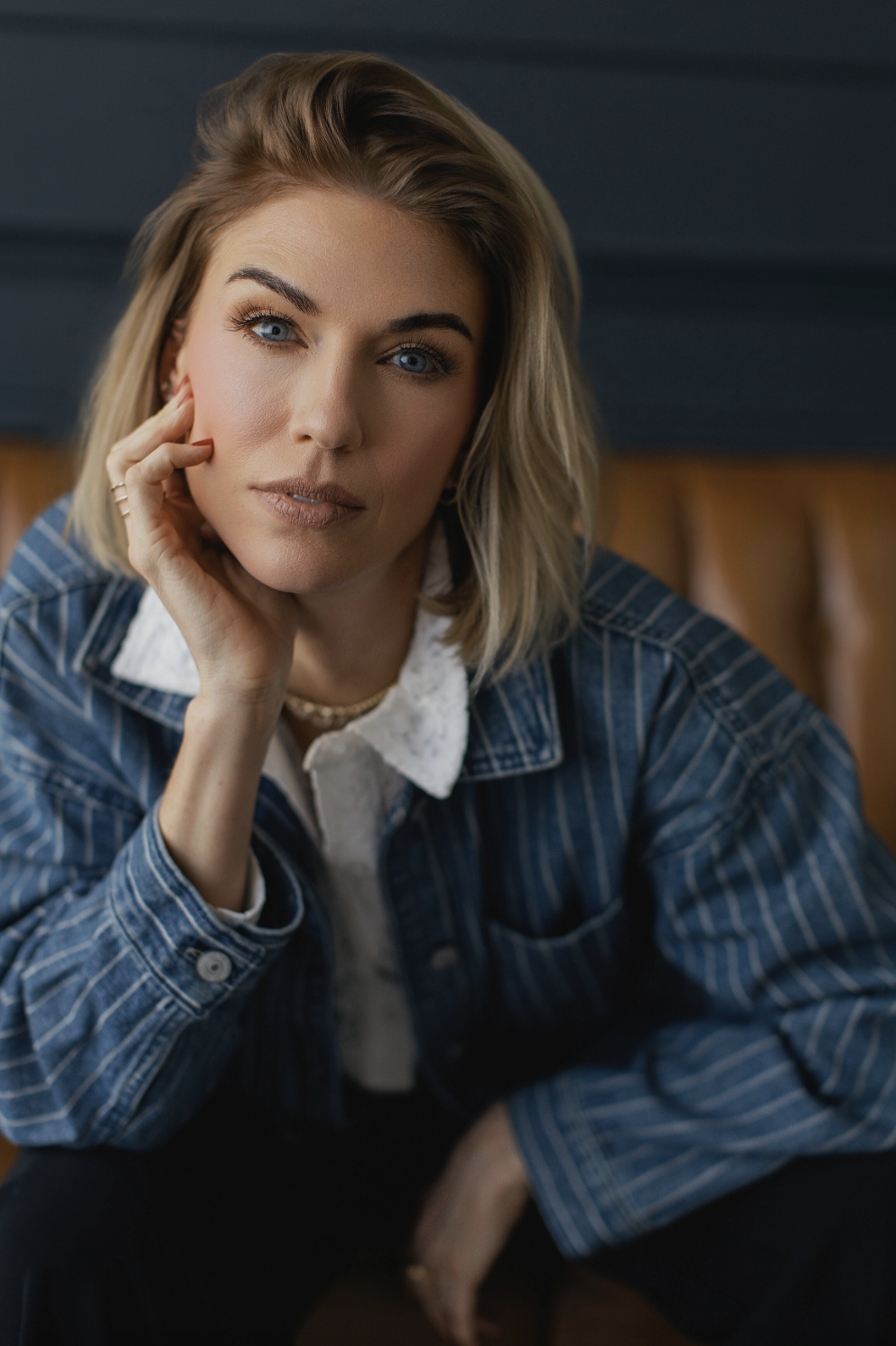 A person with blond hair and blue eyes wearing a denim jacket sits on a brown leather seat, resting their face on their hand, looking directly at the camera. An image that perfectly captures the polished yet approachable style of professional headshots Dallas clients seek.
