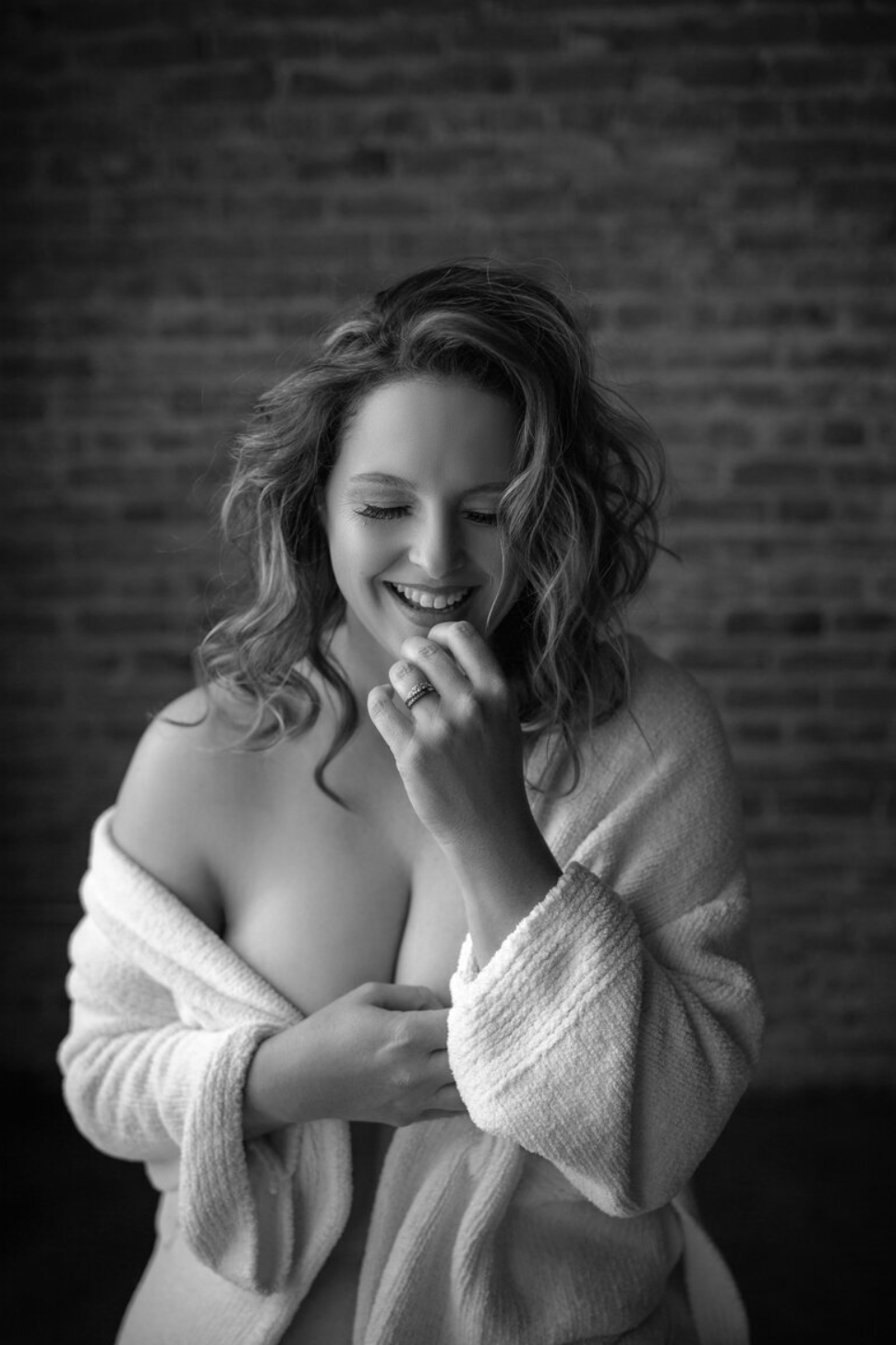 Black-and-white boudoir portrait of a smiling woman wrapped in a robe against a brick wall.