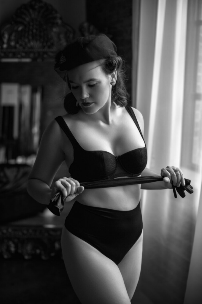 Black-and-white boudoir portrait of a woman in vintage-inspired lingerie and a hat, posing beside a window.